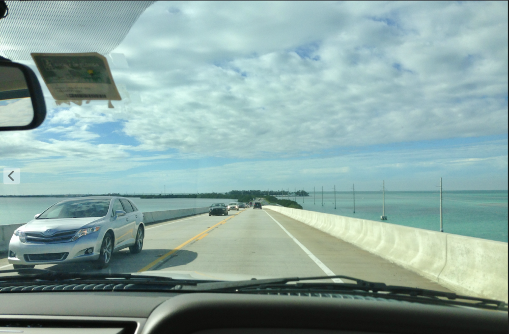 A photograph from a car dashboard of a highway over the ocean. A silver car approaches, and it is slightly cloudy overhead. 