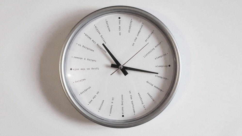 A silver clock hanging in a white wall. Instead of numbers, it has activities like "studying" "walking" "listening music" and "sleeping" listed.