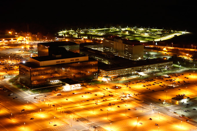 Photo of the National Security Agency in Fort Meade, Maryland. One of the Agency's buildings is dark and has mirrored glass windows, while the other to its left is low and dark, and contains another higher building rising from its center. The parking lot in front of the buildings is mostly empty. The photo is taken at night, so the orange lights of the parking lot along with white lights in a field in the back of the photo are very prominent.