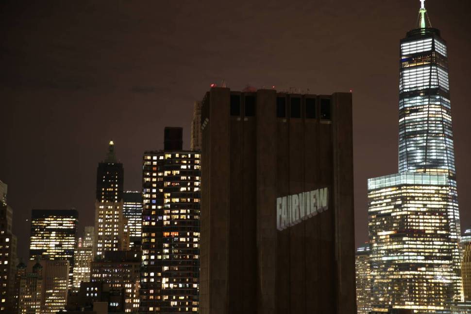 33 Thomas Street at night. It is dark, surrounded by well-lit buildings including the World Trade Center. The word "Fairview" is projected onto its surface.