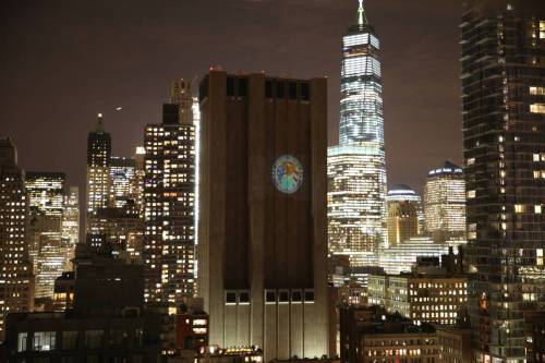 33 Thomas Street at night. It is dark, surrounded by well-lit buildings including the World Trade Center. A governmental emblem is projected onto its surface.
