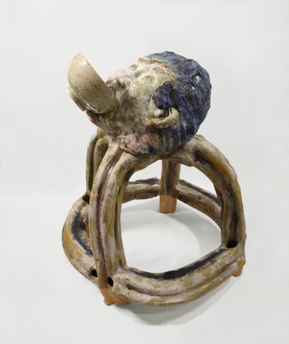 An abstract ceramic sculpture against a grey-white background. The sculpture is a stool in tones of grey, white and blue. Instead of a seat, there is the head of a man with grey skin and blue hair. He has a bowl in his mouth.