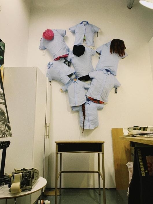 Eight short-sleeved light blue button-down shirts are stuffed and hung on a wall, in an upside-down pyramid arrangement. There are wigs and hats attached to the stuffed shirts. Below the wall arrangement is a school desk.