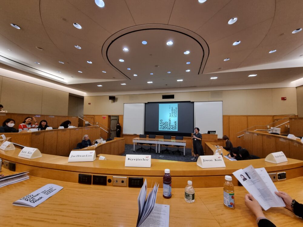 A conference style room is set up with the author's name displayed on a place card visible in the foreground. The room is filled with wooden desks and a screen against the back wall displaying the Korean Art Week poster.