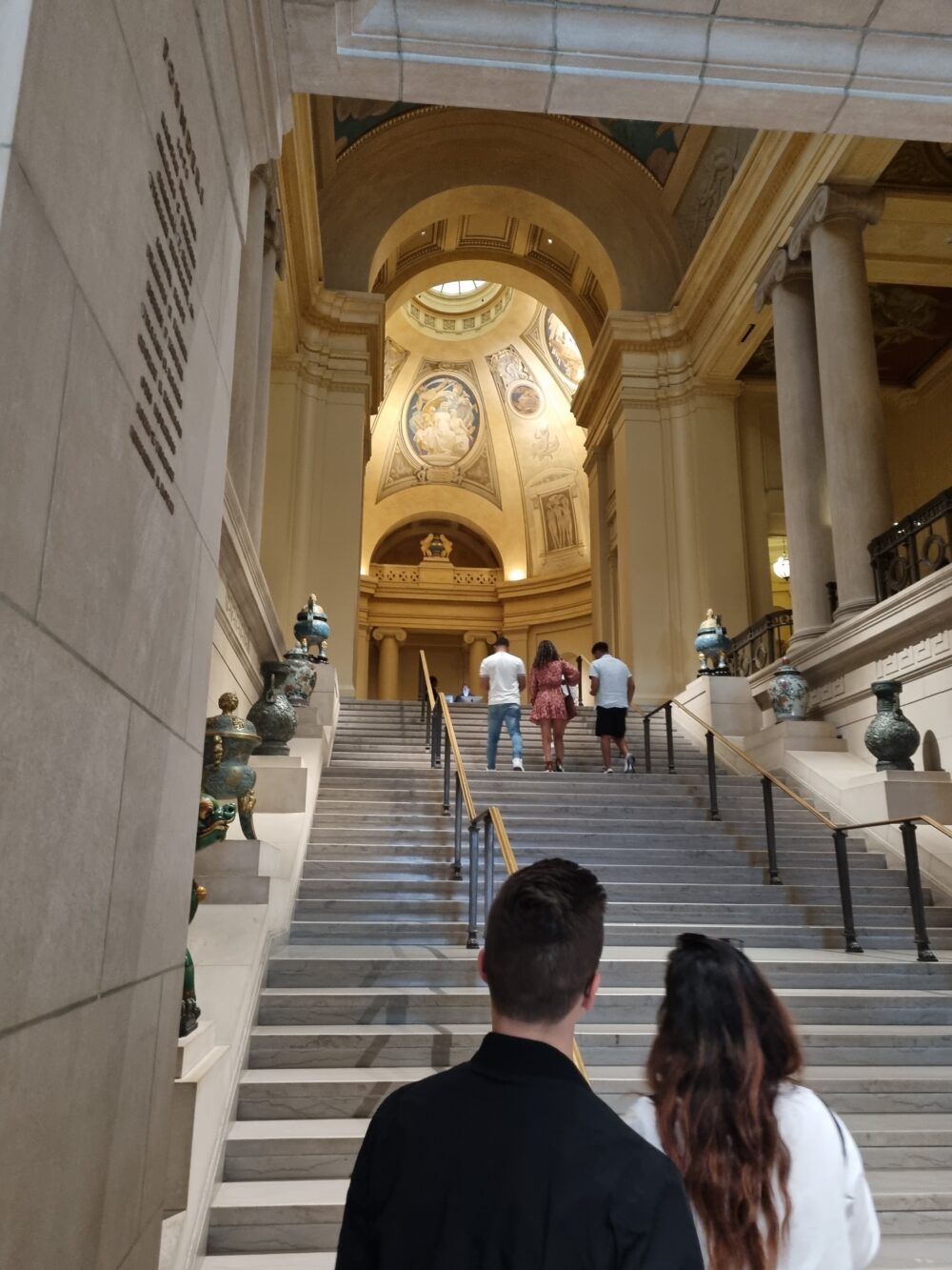Interior stairway in the Boston Museum of Fine Arts. Two individuals have their backs turned to the camera and look up the staircase which is lined with ancient vases and leads to a bright rotunda.