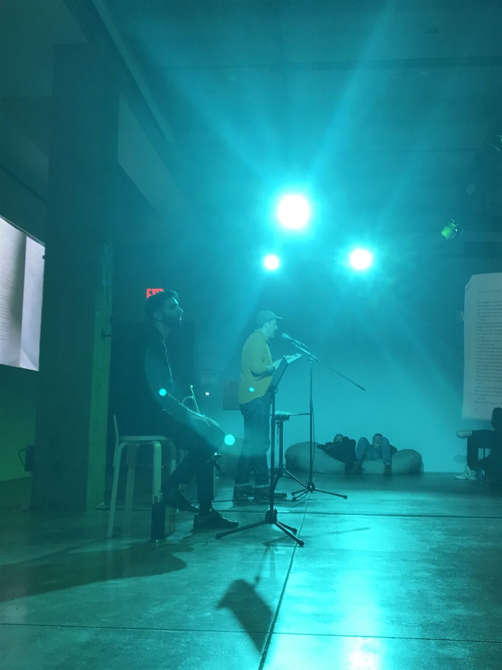 One man stands and another sits at the center of a room lit by aqua blue lights, causing the room to look as if it's underwater. The photo captures their profiles and was taken around 20 feet away. One man wears a hat and yellow shirt while the other wears dark clothes and holds a trumpet.