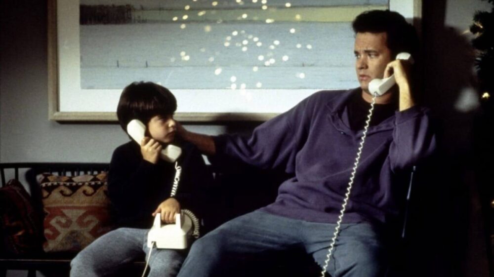 The actor Tom Hanks sits with young actor Ross Malinger on a couch, both holding white landline phones. Hanks wears a navy long-sleeved shirt and jeans. Malinger wears jeans and a black shirt. They look at each other with quizzical glances.