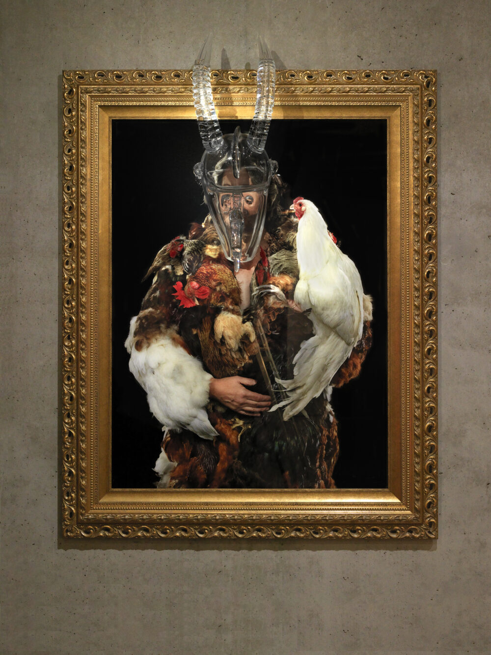 A painting of a man wearing a coat made of chicken feathers holds a white chicken in one hand. An antelope head made out of glass is placed on top of the subject's face atop the canvas. The piece is surrounded by a gold, gilded frame.