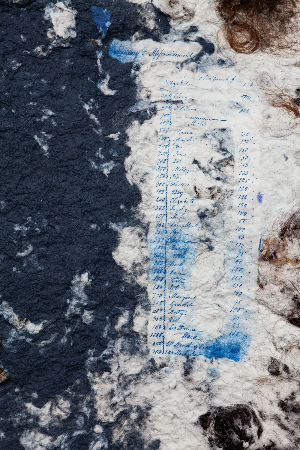 A close up image of heavily textured handmade paper with navy blue ink, human hair in the top right corner, and light blue ink depicting a list written in illegible cursive.