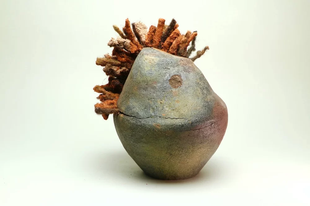 A small, mottled grey, ochre yellow, and terracotta colored clay sculpture contains various shades of orange dreadlocks sprouting from its top.