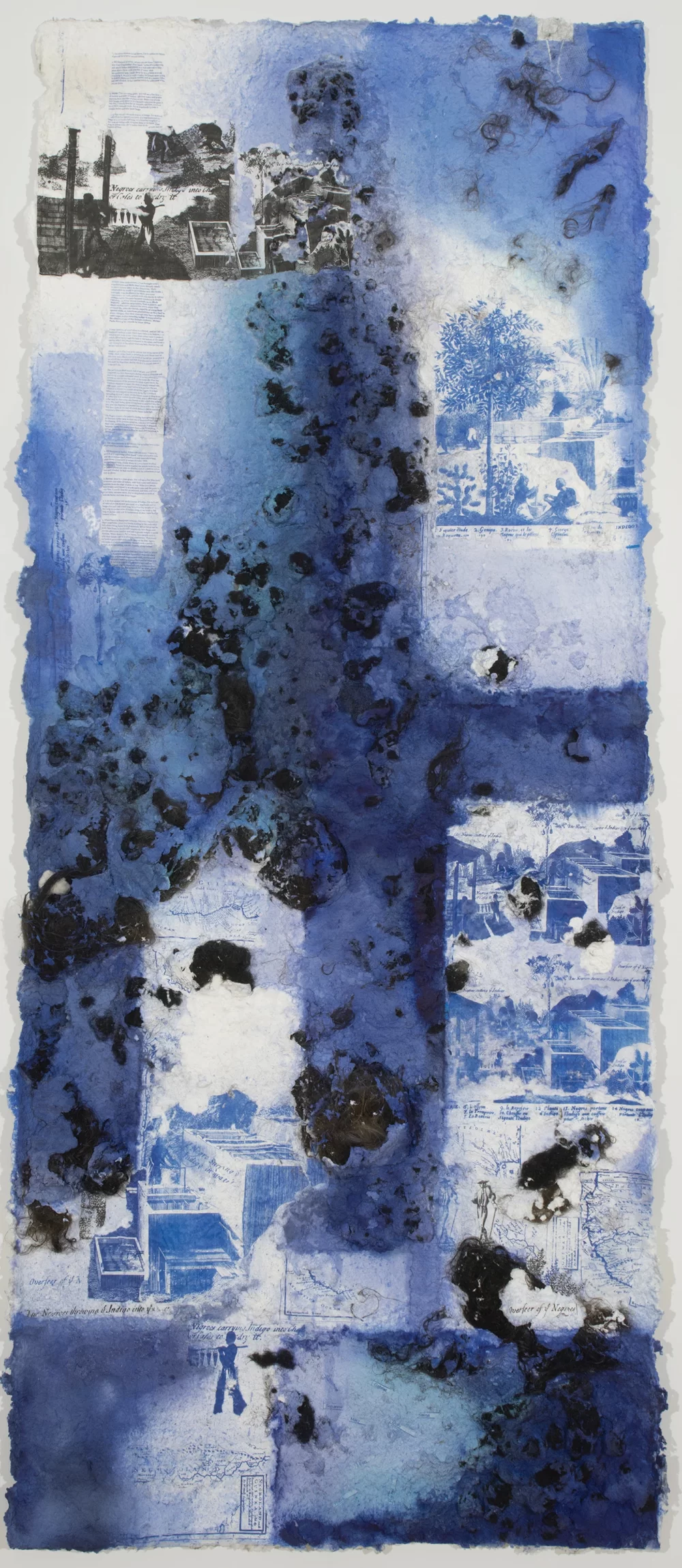 A long rectangular handmade paper panel made of hair and paper pulp and dyed various shades of blue has several printed images on it. The images show houses, a tree, and some illegible text.