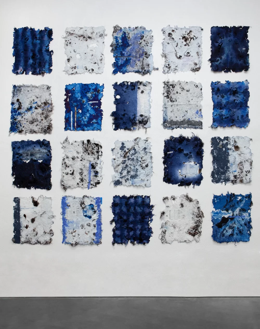 20 blue and white rectangles of hand-made paper exist as a grid on a white gallery wall. Each rectangle is different and some contain text and faint, printed imagery.