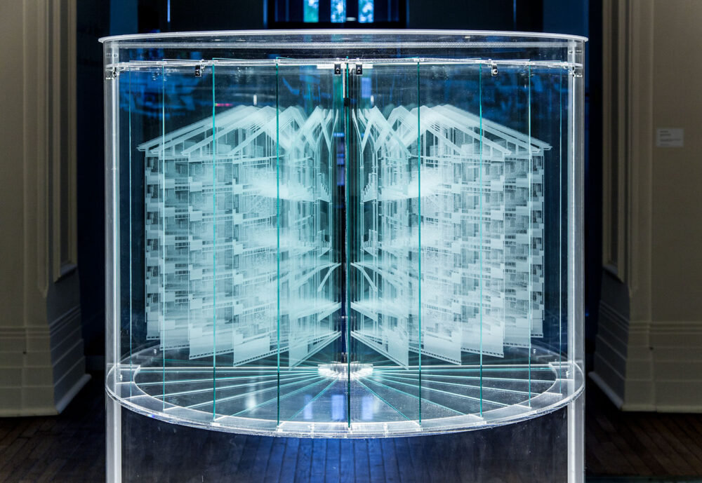 A sculpture made of carved glass based on Jeremy Bentham's panopticon prison. Panes of glass are arranged in a semi-circle from a center point, and each sheet is carved to look like a slice of a panopticon. The glass is lit so that the carvings are white, while the glass is translucent. 