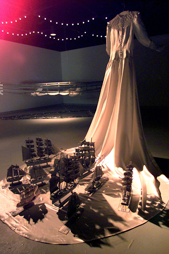 Betye Saar's Brides of Bondage, a white wedding dress hanging from the ceiling pictured from behind. The sleeves of the dress are pulled forward, and numerous miniature boats are on the train of the dress.